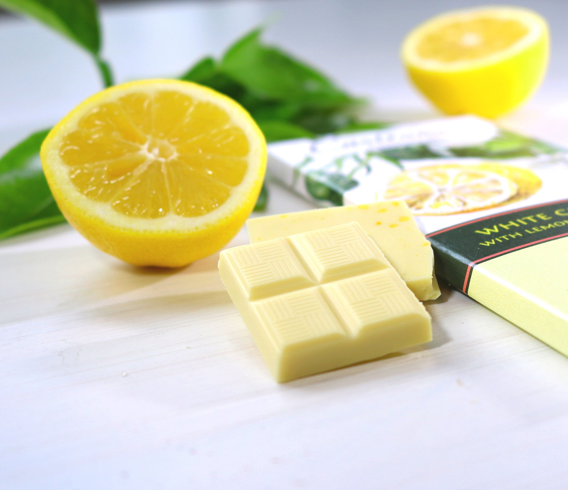 Zoomed in photo of piece of Castronovo brand white chocolate with lemon and lemon sea salt on a white cloth surface, accompanied with sliced lemon 